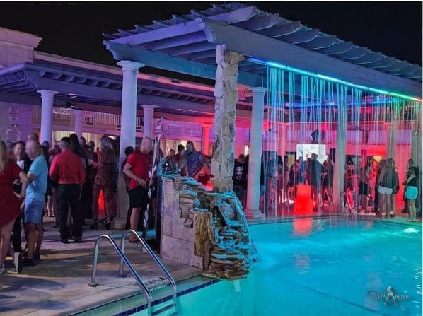 The pool deck and pergola early in a TampArotic hotel-takeover evening in Tampa Bay, with guests lit by neon at the water's edge.